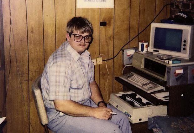 Foto dos anos 80 de um homem sentado em frente a um computador e impressora antigos.