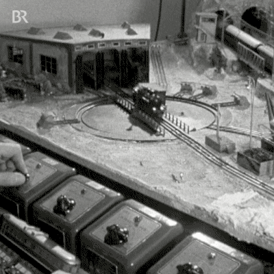 Imagem animada em preto e branco de um trem em miniatura e, ao lado, uma mão controlando em um painel de botões.